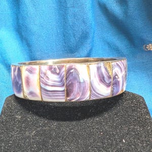 Op de afbeelding: Een brede armband bestaande uit een reeks rechthoekige segmenten van gepolijst paars en wit schelpinlegwerk. De segmenten zijn gevat in een metalen frame, wat een doorlopende band vormt met een glad, gebogen oppervlak en natuurlijke patronen.