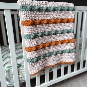 Op de afbeelding: Een roze, groene en oranje gestreepte gehaakte babydeken met een textuur bobbelsteek. De deken hangt aan een wit babybedje.