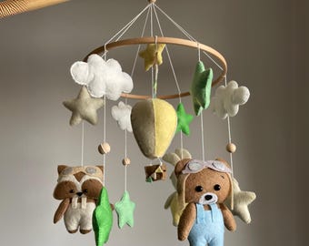 May include: A handmade baby mobile featuring felted raccoon and bear figures wearing aviator goggles and overalls. The mobile includes felt stars, clouds, a hot air balloon, and wooden beads, all suspended from a wooden hoop.