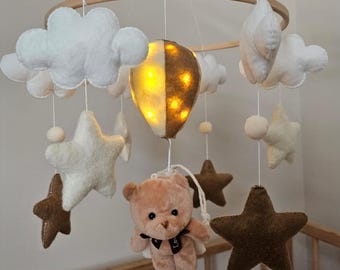 May include: A white and brown felt baby mobile with a light-up hot air balloon, clouds, stars, and a teddy bear.