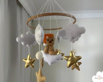 May include: A white and gold baby mobile with a teddy bear wearing a red and orange hat, clouds, and stars. The mobile is hanging from a wooden ring.