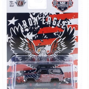 May include: A die-cast model of a 1974 Chevrolet K5 Blazer, featuring a patriotic American flag design. The model is packaged in a clear plastic case with a backdrop of the American flag and the words "Iron Eagle".