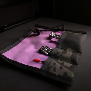 May include: A roll-up watch storage pouch featuring a vibrant purple interior and a dark camouflage exterior. The pouch includes multiple slots for timepieces, metal watch buckles, and a small tool with a bright red tip, secured by a black cord.