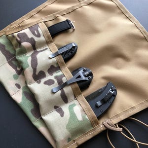 May include: A tan fabric roll-up organiser featuring a camouflage patterned pocket section. The organiser contains three black folding knives and a black watch strap, each secured within individual vertical slots. The edges are stitched.