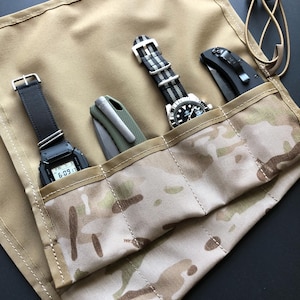May include: A tan fabric gear roll with a camouflage pattern pocket section containing a digital watch with a black strap, a folding knife with a green handle, an analogue watch with a striped strap, and a black folding knife with a pocket clip.
