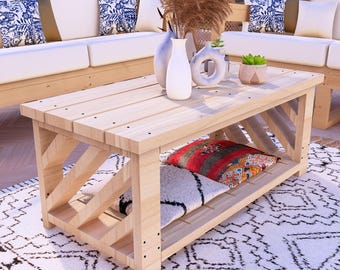 Puede incluir: Una mesa de centro de madera de color claro con un estante inferior que contiene dos cojines decorativos. La mesa tiene un soporte en forma de X en cada extremo. Sofás blancos con cojines estampados azules en el fondo.