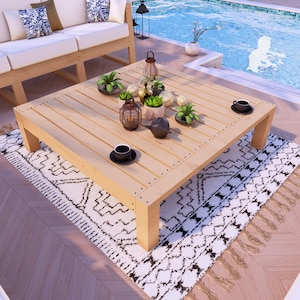 May include: A square wooden coffee table with a natural wood finish. The table is surrounded by a white and black patterned rug with fringe. There are several small potted plants and a teapot on the table.