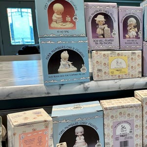May include: A collection of blue and white boxes with Precious Moments figurines. The boxes are stacked on a shelf and each box features a different figurine and inscription. The inscriptions include "In His Time", "You Will Always Be My Choice", "You Are A Blessing To Me", "God Bless You For Touching My Life", and "Grandma's Prayer".