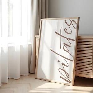 May include: A framed print with the word "Pilates" in a brown script font. The print is in a light wooden frame and leans against a wall near a window with sheer curtains. The background includes a wooden cabinet.