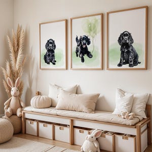 Op de afbeelding: Drie ingelijste aquarellen van zwarte cocker spaniel puppy's. De schilderijen hangen aan een lichtbeige muur boven een witte bank met beige kussens. De bank heeft vier opbergladen.