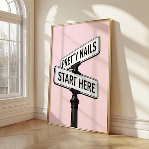 May include: Framed print with a pink backdrop, displaying two street signs. The top sign reads "PRETTY NAILS", the bottom "START HERE". The signs are white with black text, mounted on a black post. The frame is gold-colored.