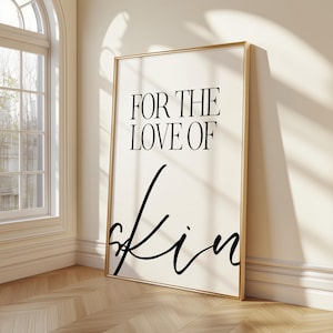 May include: A framed art print with the words "FOR THE LOVE OF Skin" in black script and serif fonts. The print has a cream-colored background and a gold frame. The artwork is leaning against a wall in a room with a window.