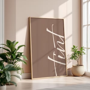 May include: A framed art print with the word "Tint" in white script against a brown background. The artwork is leaning against a wall, with potted plants in the foreground and a window in the background. The frame is a light wood color.