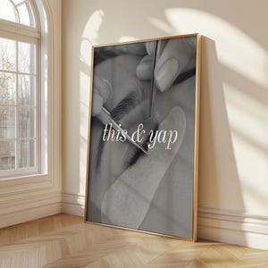 Op de afbeelding: Een zwart-wit ingelijste wandkunstprint met een close-up van een wimperextensions-applicatie met metalen pincetten en de witte tekst this & yap gecentreerd over de afbeelding. De print zit in een dunne gouden metalen lijst.