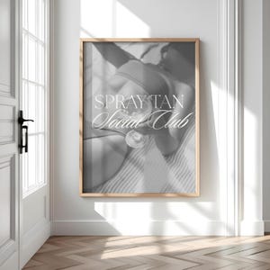 May include: Monochrome framed print with the text "SPRAY TAN Social Club". The image shows a person holding a glass of liquid. The print is displayed on a white wall with a wooden frame.
