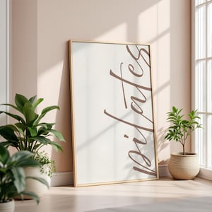 May include: A framed print with the word "Pilates" in a brown script font. The frame is gold-colored, and the background is off-white. The print is leaning against a wall, with potted plants in the foreground.