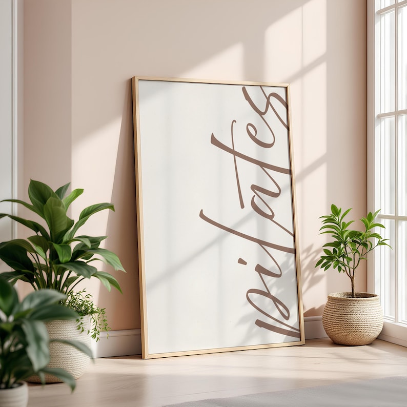May include: A framed print with the word "Pilates" in a brown script font. The frame is gold-colored, and the background is off-white. The print is leaning against a wall, with potted plants in the foreground.