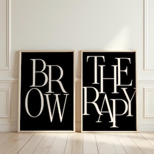 Op de afbeelding: Twee ingelijste kunstprints met de woorden "BROW" en "THERAPY" in een witte schreefletter op een zwarte achtergrond. De lijsten zijn licht van kleur en de prints worden getoond op een witte muur boven een houten vloer.