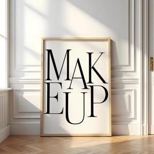 Könnte beinhalten: Gerahmter Druck mit dem Wort "MAKEUP" in fetten, schwarzen Großbuchstaben. Das Kunstwerk wird auf weißem Hintergrund, in hellem Holz gerahmt, vor einer weißen Wand und einem Holzboden gezeigt.