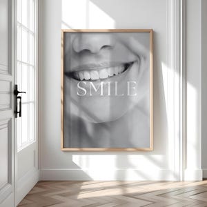 以下が含まれることがあります： 白い文字で「SMILE」という言葉が入った、笑顔の口のクローズアップを特徴とする、額装された白黒プリント。プリントは明るい木製のフレームで、白い壁に掛けられています。画像は木の床がある部屋にあります。