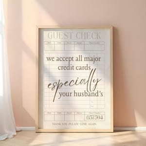 May include: A framed guest check print with a beige background and a wooden frame. The text reads "GUEST CHECK" at the top, followed by "we accept all major credit cards especially your husband's". The check number is 037594.