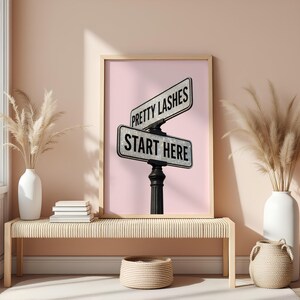 May include: A framed print featuring a pink background with two street signs that read "Pretty Lashes" and "Start Here." The signs are mounted on a black post. The print is displayed above a woven bench with decorative vases.