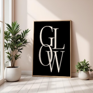 Puede incluir: Un cuadro enmarcado con la palabra "GLOW" en letras blancas grandes y elegantes sobre un fondo negro. El marco es de color madera clara. La impresión se muestra sobre un suelo de madera blanca, con una gran planta en maceta a la izquierda y otra más pequeña a la derecha.
