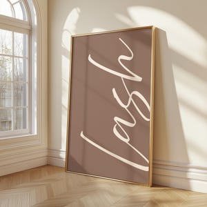 May include: A rectangular wall art print in a thin gold frame featuring a solid muted brown background with the word lash written in large, flowing, off-white cursive script lettering oriented vertically across the center of the print.