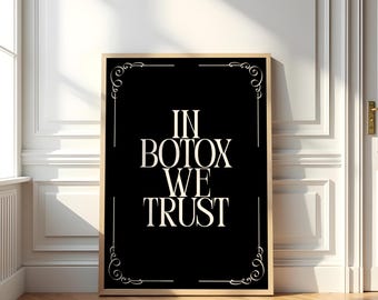 May include: A framed art print with the text "IN BOTOX WE TRUST" in white, centered on a black background. The print has a gold-colored frame and decorative white corner accents. The artwork is displayed on a wooden floor.