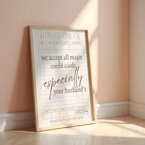 May include: A framed "Guest Check" sign with a light wood frame. The sign has a grid layout with text that reads "we accept all major credit cards, especially your husband's." The check number is 037594.