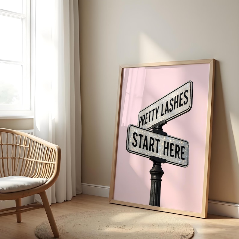 May include: A framed print featuring a street sign design with the text "PRETTY LASHES" above "START HERE". The signs are silver with black text, set against a pink background. The frame is light brown.
