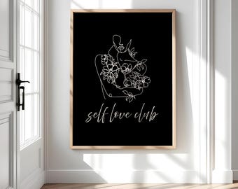 May include: A framed art print featuring a minimalist line drawing of a person embracing flowers, set against a black background. The words "self love club" are written in cursive script below the illustration. The frame is a light wood color.