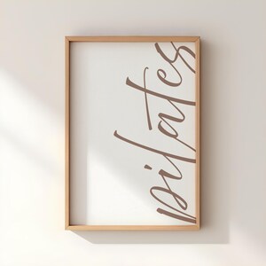May include: A framed print with the word "pilates" in a brown script font on a white background. The frame is made of light-colored wood. The print is displayed against a white wall.