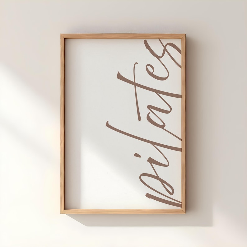 May include: A framed print with the word "pilates" in a brown script font on a white background. The frame is made of light-colored wood. The print is displayed against a white wall.