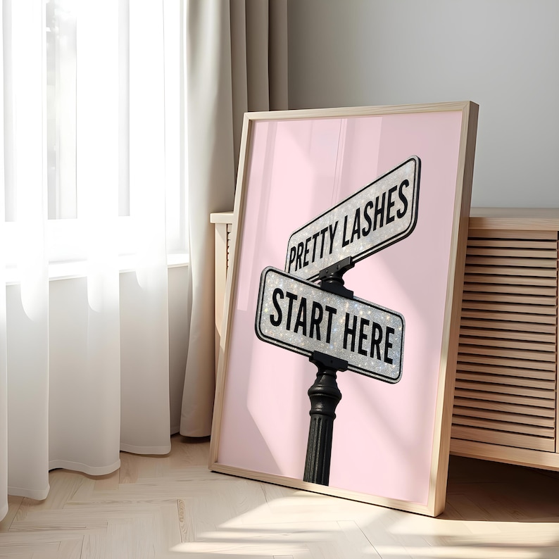 May include: A framed print featuring a pink background with two street signs. The top sign reads "PRETTY LASHES" and the bottom sign reads "START HERE" in black text. The signs are mounted on a black post.