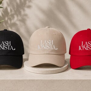 Puede incluir: Tres gorras de béisbol en negro, beige y rojo, cada una con el texto bordado en blanco LASH IONISTA en la parte frontal. Las gorras tienen un diseño estructurado con una visera curva y un botón en la parte superior, mostrando marca uniforme.