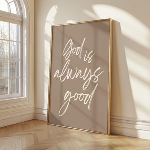 Pode incluir: Uma impressão de arte de parede retangular numa moldura dourada fina com um fundo taupe e o texto manuscrito em branco God is always good. As letras estão num estilo de caligrafia casual, centradas na impressão vertical com linhas.