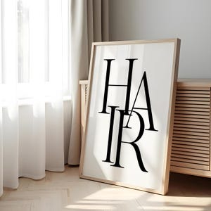 Può includere: Una stampa incorniciata con la parola "HAIR" in grandi lettere nere e in grassetto. L'opera d'arte è presentata su uno sfondo bianco all'interno di una cornice in legno chiaro. La stampa è appoggiata a un muro vicino a una finestra con tende trasparenti.