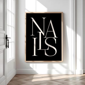 May include: A framed art print featuring the word "NAILS" in a bold, white, stylized font against a black background. The artwork is in a light wood frame and hangs on a white wall, with a door and sunlight visible.