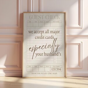 May include: A framed guest check print with a beige background and a gold frame. The text reads "GUEST CHECK" at the top, followed by "we accept all major credit cards, especially your husband's." The check number is 037594.