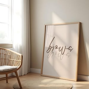 Op de afbeelding: Een ingelijste print met het woord "brows" in een stijlvol handschrift. Het kunstwerk zit in een lichte houten lijst, tegen een beige muur. Een rieten stoel en een rond tapijt staan op de voorgrond, wat een gezellige, neutraal getinte interieurscène creëert.