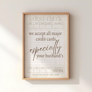 May include: A framed guest check print with a light wood frame. The text reads "GUEST CHECK" at the top, followed by lines for details. The print humorously states "we accept all major credit cards, especially your husband's." The check number is 037594.