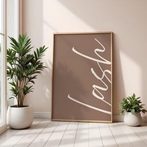 May include: A framed print with the word "lash" in white script on a brown background. The artwork is leaning against a wall, next to a potted plant and a window. The frame is a light wood colour.