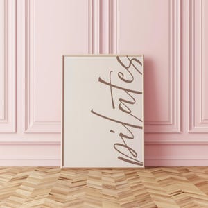 May include: A framed print with the word "pilates" in a stylish, cursive font. The print has a neutral background and a light brown frame. The background is a light pink wall with decorative paneling, and the floor is wood.