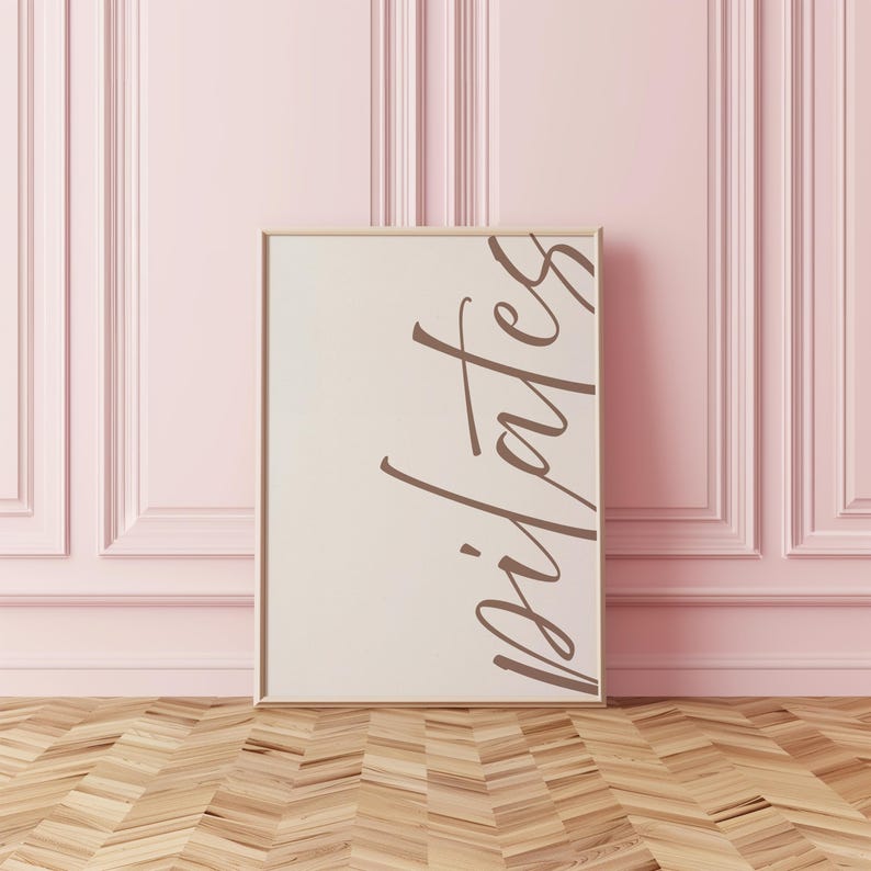 May include: A framed print with the word "pilates" in a stylish, cursive font. The print has a neutral background and a light brown frame. The background is a light pink wall with decorative paneling, and the floor is wood.