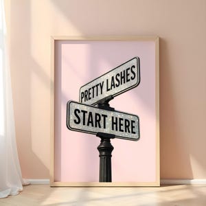 May include: Framed print featuring a pink background with a black street sign post. The sign reads "PRETTY LASHES" and "START HERE" in white text. The signs have a glittery texture, and the frame is a light wood color.