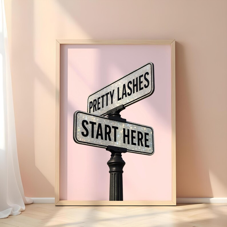 May include: Framed print featuring a pink background with a black street sign post. The sign reads "PRETTY LASHES" and "START HERE" in white text. The signs have a glittery texture, and the frame is a light wood color.