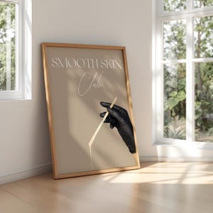 Op de afbeelding: Een ingelijst print met een beige achtergrond en de tekst "SMOOTH SKIN Club" in wit. Een hand met een handschoen houdt een houten stok vast met een witte substantie die er vanaf druppelt.
