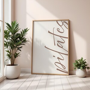 May include: A framed Pilates art print with a light wood frame. The print features the word "Pilates" in a stylish, brown script against a white background. A potted plant and a small green plant in a white pot are in the foreground.