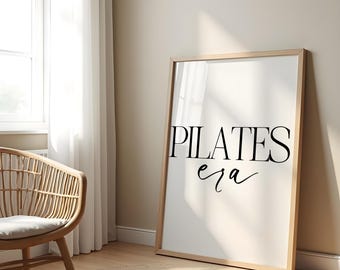 May include: A framed print with the words "PILATES era" in black script. The print is leaning against a wall in a room with a window, a woven chair, and a round rug. The frame is light-colored wood.
